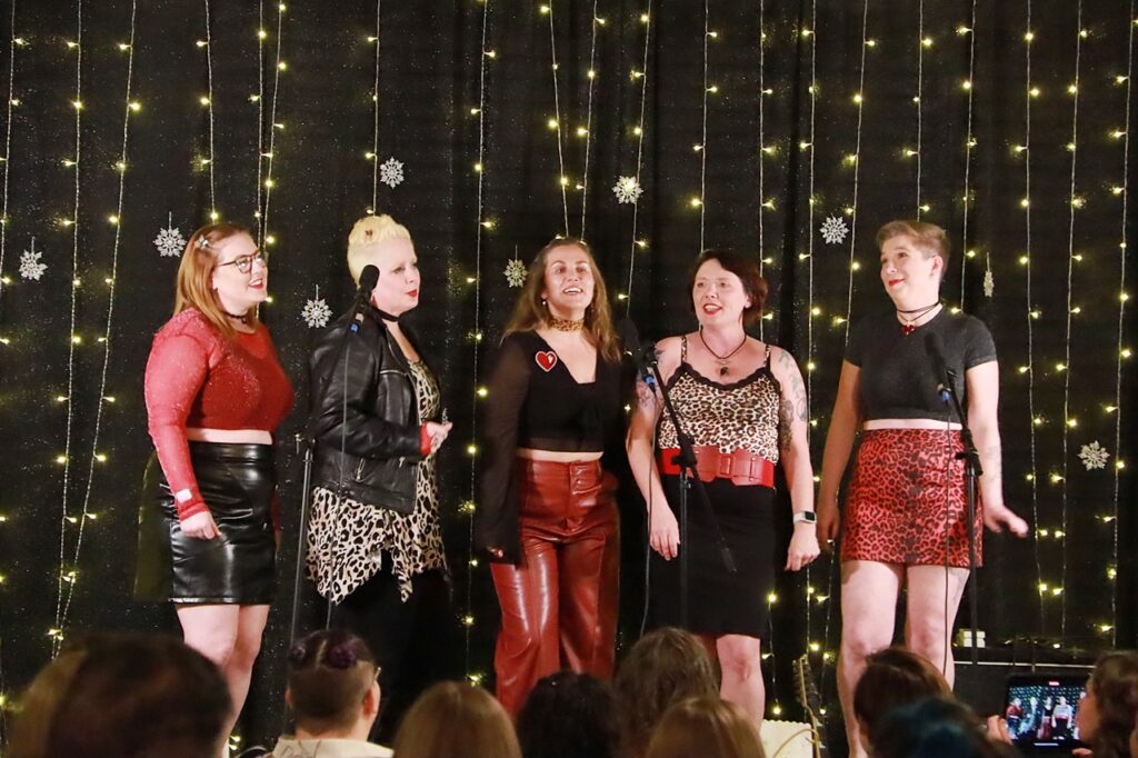 Five members of SDWC, dressed in red, black, and leopard print, sing in front of a crowd with a fairy light backdrop over a black curtain
